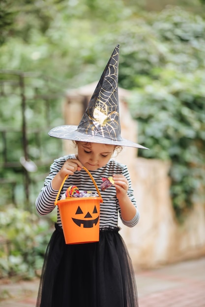 Bambina carina in costume da strega che tiene secchio di zucca jackolantern con caramelle e dolci Dolcetto o scherzetto per bambini durante le vacanze di Halloween