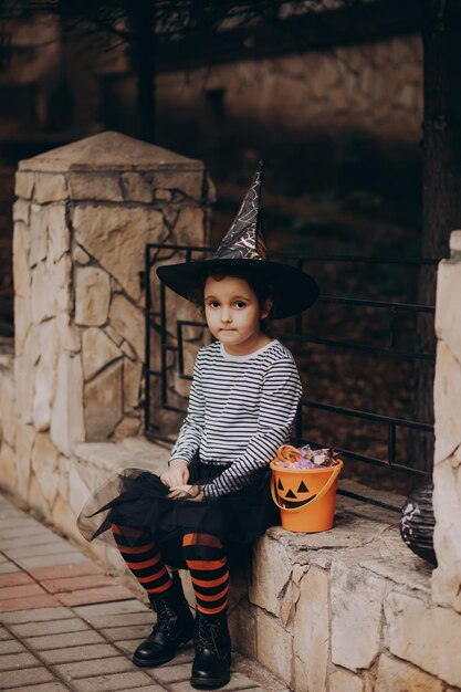 Bambina carina in costume da strega che tiene secchio di zucca jackolantern con caramelle e dolci Dolcetto o scherzetto per bambini durante le vacanze di Halloween