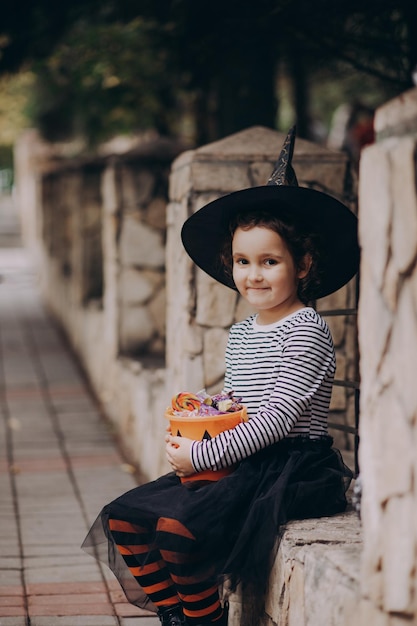 Bambina carina in costume da strega che tiene secchio di zucca jackolantern con caramelle e dolci Dolcetto o scherzetto per bambini durante le vacanze di Halloween