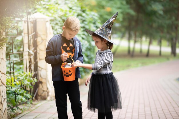 Bambina carina in costume da strega e ragazzo che tiene secchio di zucca jack-o-lantern con caramelle e dolci e caramelle ai gusti. Dolcetto o scherzetto per bambini durante le vacanze di Halloween.