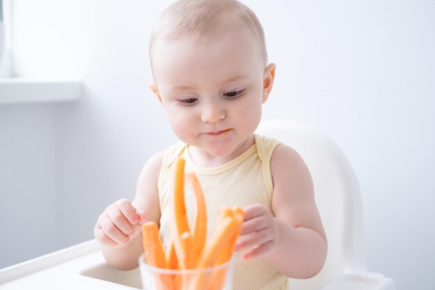 bambina carina in tuta gialla seduta sulla sedia del bambino che mangia fette di carota