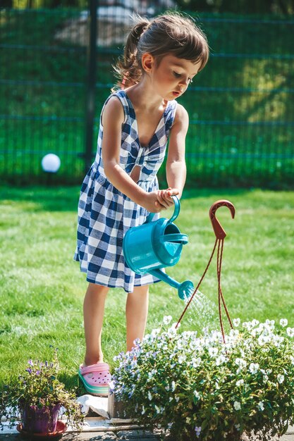 Bambina carina in un vestito scozzese blu che innaffia le piante da un annaffiatoio in giardino