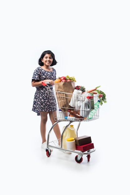 Bambina carina indiana con carrello della spesa o carrello pieno di generi alimentari, frutta e verdura, isolato su muro bianco