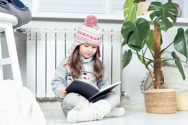 Bambina carina seria interessata in cappello di lana e vestiti caldi con libro di lettura del pupazzo di neve accanto al radiatore di riscaldamento