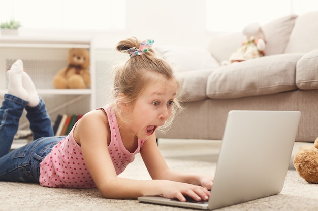 Bambina casuale spaventata che guarda film sul computer portatile mentre è sdraiata sul pavimento a casa