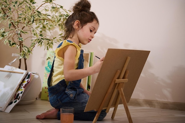 Bambina caucasica allegra e carina seduta su cavalletto in legno e pittura su tela Concetto di educazione artistica e creatività per l'intrattenimento per bambini
