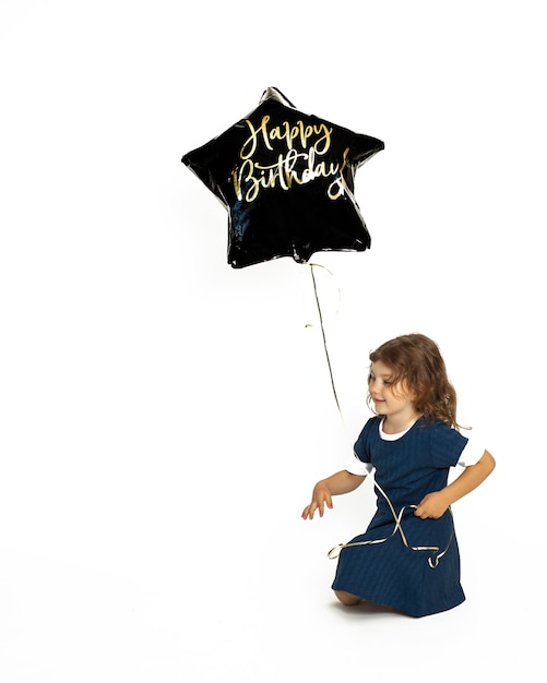Bambina caucasica carina 4-5 anni seduta felicemente con palloncino di gel nero con scritta Happy Birthday. Foto in studio su sfondo bianco
