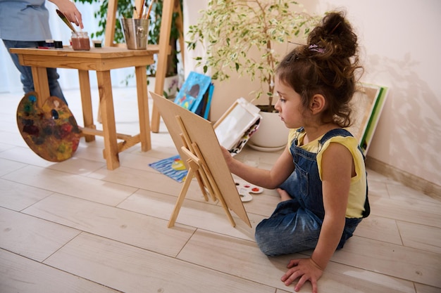 Bambina caucasica concentrata che si diverte a dipingere seduta sul pavimento su un cavalletto di legno sullo sfondo di bambini gioiosi colorati dipinti immagini Creatività e istruzione di classe artistica