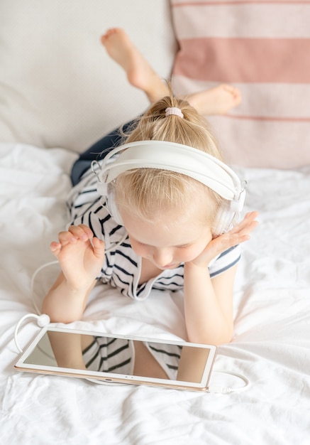 Bambina caucasica in cuffia che guarda il tablet a letto, interni domestici, moderne tecnologie dei dispositivi stile scandinavo