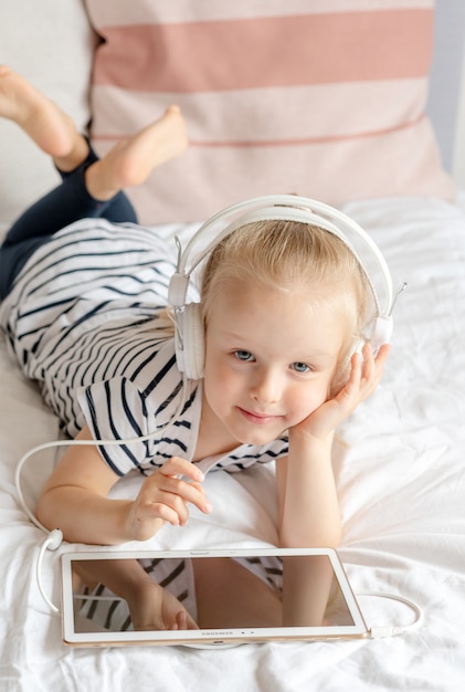Bambina caucasica in cuffia che guarda il tablet a letto, interni domestici, moderne tecnologie dei dispositivi stile scandinavo