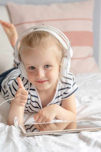 Bambina caucasica in cuffia che guarda il tablet a letto, interni domestici, moderne tecnologie dei dispositivi stile scandinavo