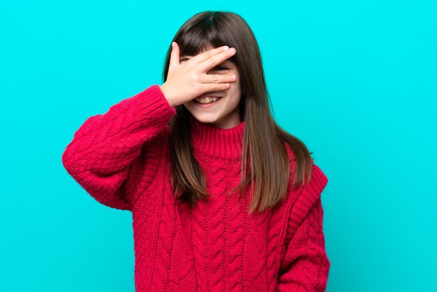 Bambina caucasica isolata su sfondo blu che copre gli occhi con le mani e sorridente