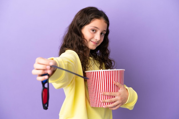 Bambina caucasica isolata su sfondo viola con occhiali 3d e in possesso di un grande secchio di popcorn