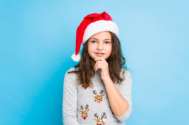 Bambina che celebra il giorno di Natale sorridendo felice e fiducioso, toccando il mento con la mano.