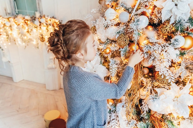 Bambina che decora l'albero di Natale alla vigilia di Natale a casa. Ragazzino in camera da letto leggera con decorazioni invernali. Famiglia felice a casa. Natale Capodanno dicembre per il concetto di celebrazione.