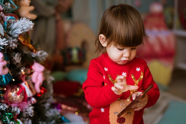 Bambina che gioca al cellulare con un maglione natalizio rosso accanto a bambini dell'albero fes...