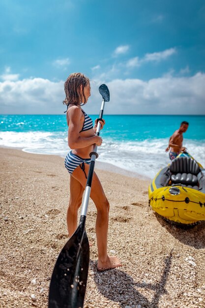 Bambina che gioca con una pagaia accanto a un kayak che suo padre prepara per la navigazione in mare.