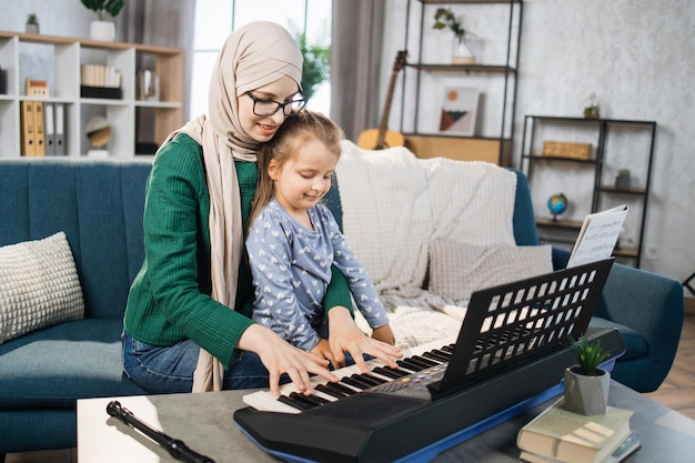 Bambina che impara a suonare il pianoforte usando le note insieme a sua madre musulmana in hijab