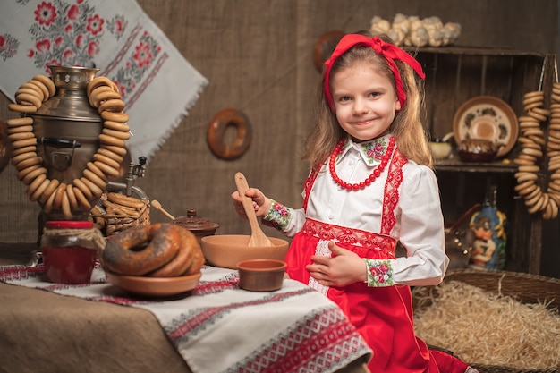 Bambina che indossa la fascia rossa e camicia ornamentale seduto al tavolo pieno di cibo e grande samovar celebrando Maslenitsa