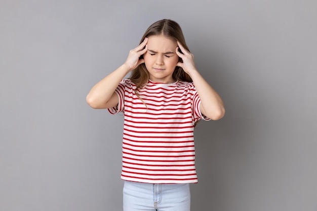 Bambina che indossa una maglietta a righe accigliata stringendo la testa dolorante che soffre di intenso mal di testa