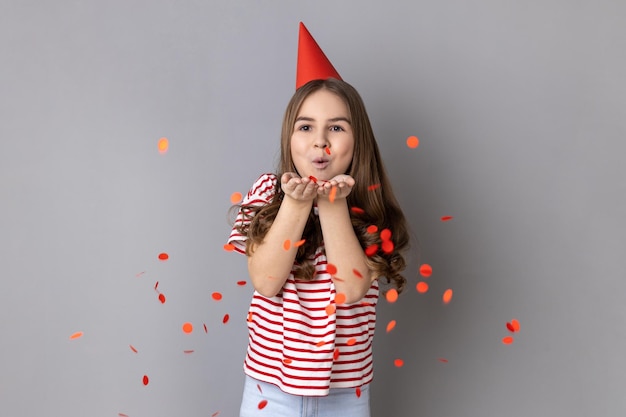 Bambina che indossa una maglietta a righe e un cono di festa che soffia coriandoli a forma di cuore godendosi il compleanno