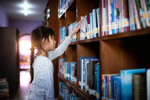 Bambina che sceglie il libro nella sala della biblioteca pubblica selezionando la letteratura per la lettura la ragazza sceglie il libro