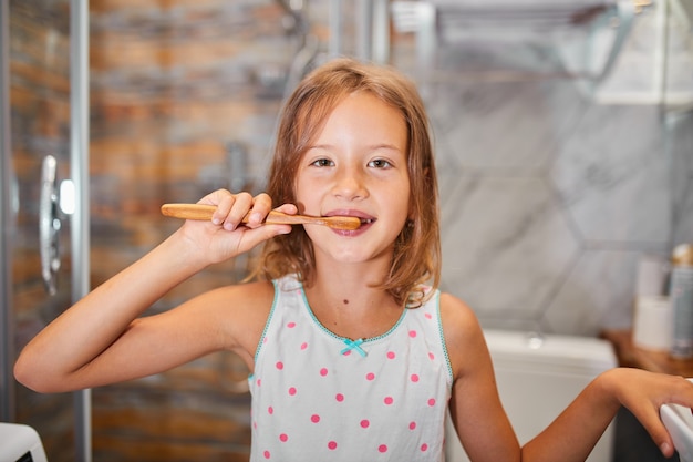 Bambina che si lava i denti con lo spazzolino da denti in legno ecologe nel bagno di casa, concetto di igiene sana