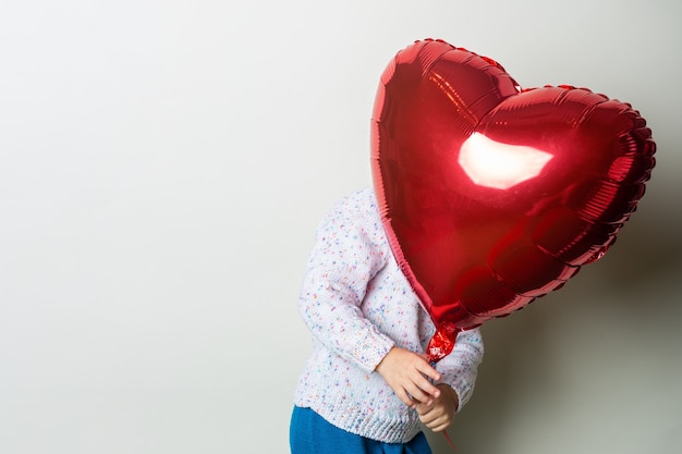 Bambina che si nasconde dietro una mongolfiera cuore su sfondo chiaro. Concetto per il giorno di San Valentino, compleanno. Banner.