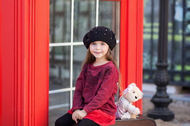 Bambina che si siede su una valigia con un orsacchiotto. Cabina telefonica rossa di Londra. Primavera. Autunno. Con la giornata internazionale della donna. Dall'8 marzo! Ritratto del primo piano del bambino della bambina del fronte.