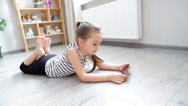 Bambina che si trova su un pavimento di legno con un telefono, guardando un cartone animato o facendo una videochiamata