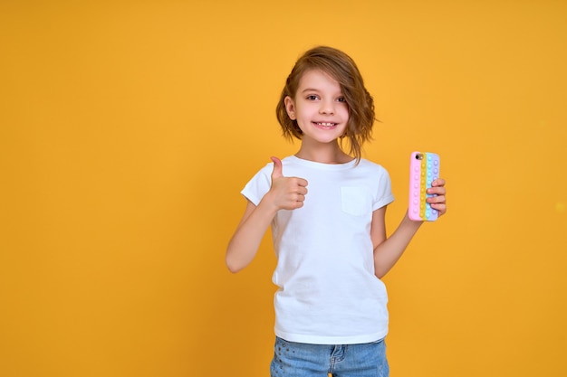 Bambina che tiene il telefono e mostra il pollice in alto isolato su uno sfondo di colore arancione