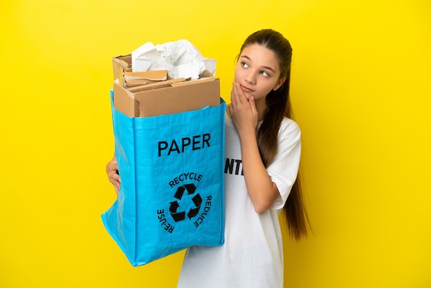 Bambina che tiene in mano un sacchetto per il riciclaggio pieno di carta da riciclare su una superficie gialla isolata che guarda in alto sorridendo