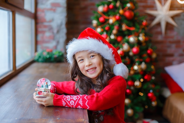 Bambina che tiene un globo di neve di vetro sullo sfondo del capodanno di Natale di un albero di Natale