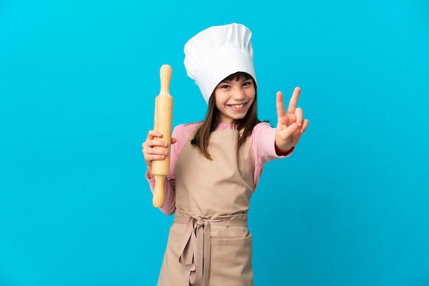 Bambina che tiene un mattarello isolato su sfondo blu sorridendo e mostrando il segno di vittoria