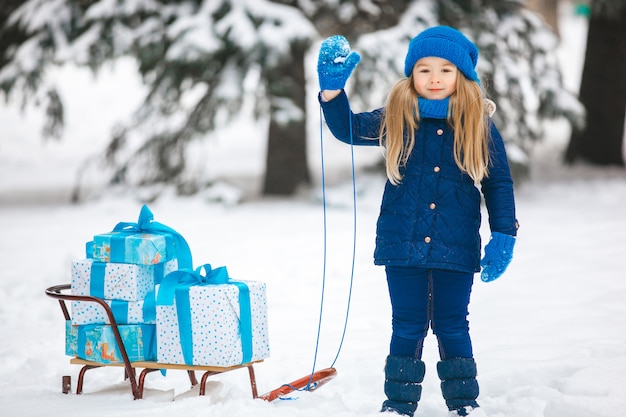 Bambina che tiene un regalo di natale in sue mani e che sorride emotivamente. Bambino che indossa un cappello blu a maglia, guanti, guanti, sciarpa