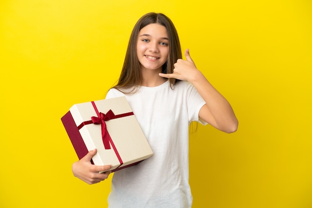 Bambina che tiene un regalo sopra fondo giallo isolato che fa il gesto del telefono. Richiamami segno