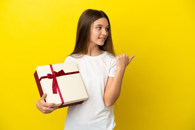 Bambina che tiene un regalo su sfondo giallo isolato che punta al lato per presentare un prodotto