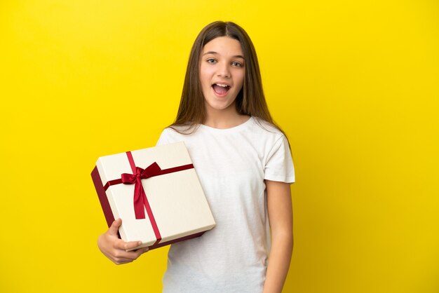 Bambina che tiene un regalo su sfondo giallo isolato con espressione facciale a sorpresa