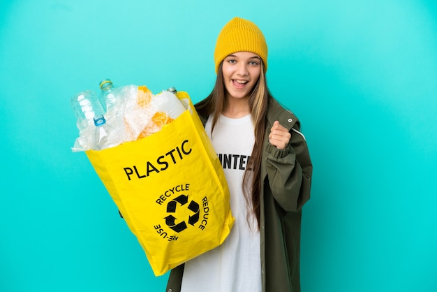 Bambina che tiene una borsa piena di bottiglie di plastica da riciclare su sfondo blu isolato che celebra una vittoria nella posizione del vincitore