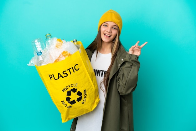 Bambina che tiene una borsa piena di bottiglie di plastica da riciclare su sfondo blu isolato che sorride e mostra il segno della vittoria