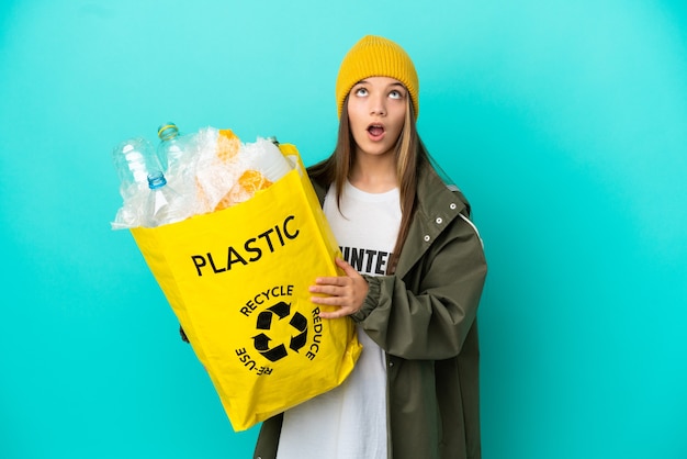 Bambina che tiene una borsa piena di bottiglie di plastica da riciclare su sfondo blu isolato guardando in alto e con espressione sorpresa