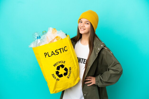 Bambina che tiene una borsa piena di bottiglie di plastica da riciclare su un muro blu isolato in posa con le braccia all'anca e sorridente