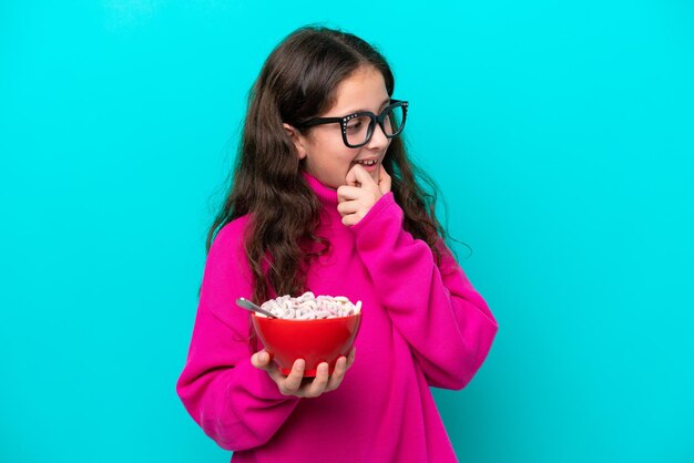 Bambina che tiene una ciotola di cereali isolata su sfondo blu pensando un'idea e guardando il lato