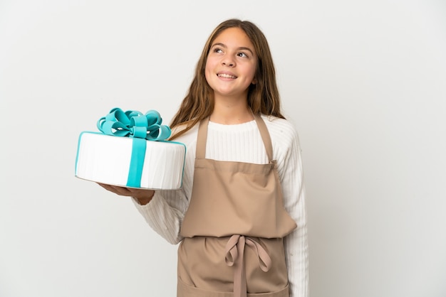 Bambina che tiene una grande torta sopra fondo bianco isolato che pensa un'idea mentre osserva in su