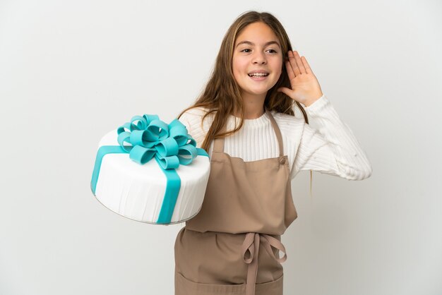 Bambina che tiene una grande torta su sfondo bianco isolato ascoltando qualcosa mettendo la mano sull'orecchio