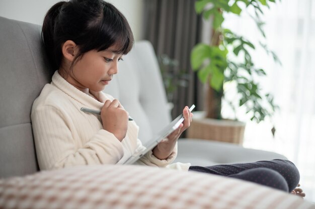 Bambina che utilizza tablet per giocare su Internet Bambino seduto sul divano a guardare o parlare con un amico online Bambino che si rilassa in soggiorno al mattino Bambini con il nuovo concetto di tecnologia