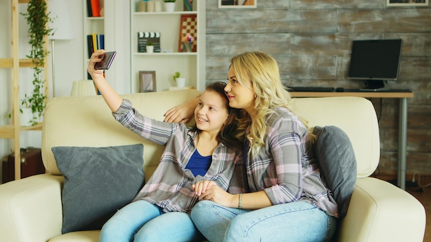 Bambina con bretelle seduta sul divano in soggiorno prendendo un selfie con sua madre.