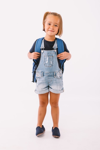 Bambina con capelli biondi vestita con una salopette blu e una t-shirt, con uno zaino pronto per il ritorno a scuola, su sfondo bianco