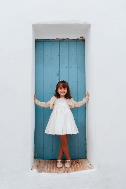 Bambina con capelli castani appoggiata a una porta blu in un vestito