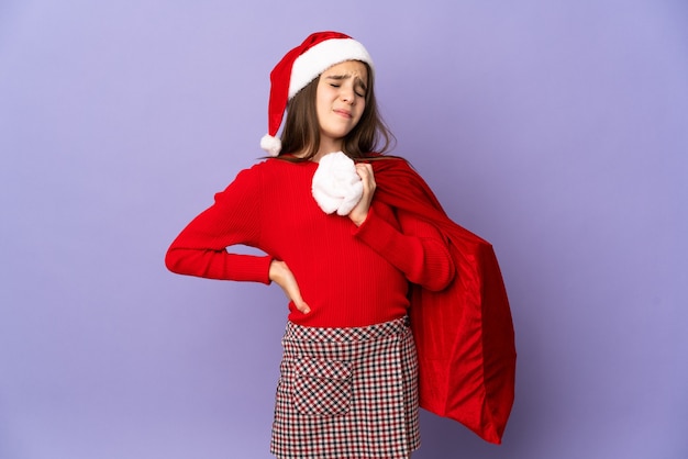 Bambina con cappello e sacco di Natale isolato su sfondo viola soffre di mal di schiena per aver fatto uno sforzo